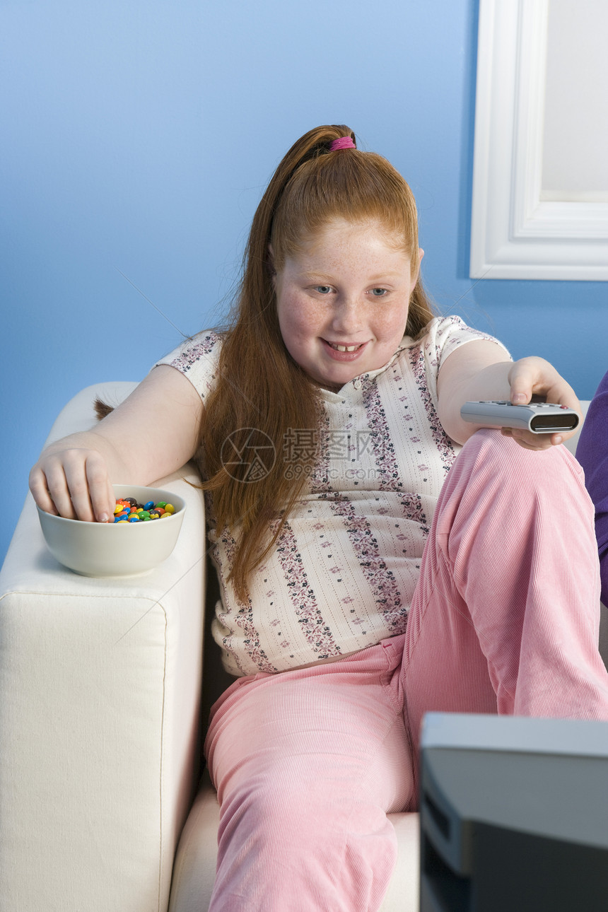 一个带着遥控器的笑笑超重女孩的肖像 在沙发上吃甜食图片