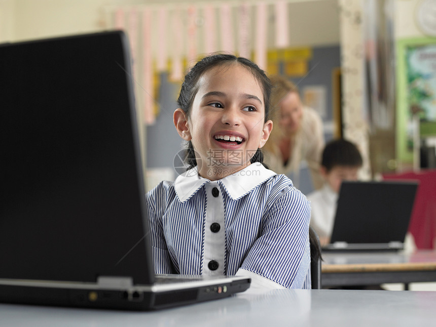 带着教师和背景男孩坐在笔记本电脑上时微笑的年轻女学生图片