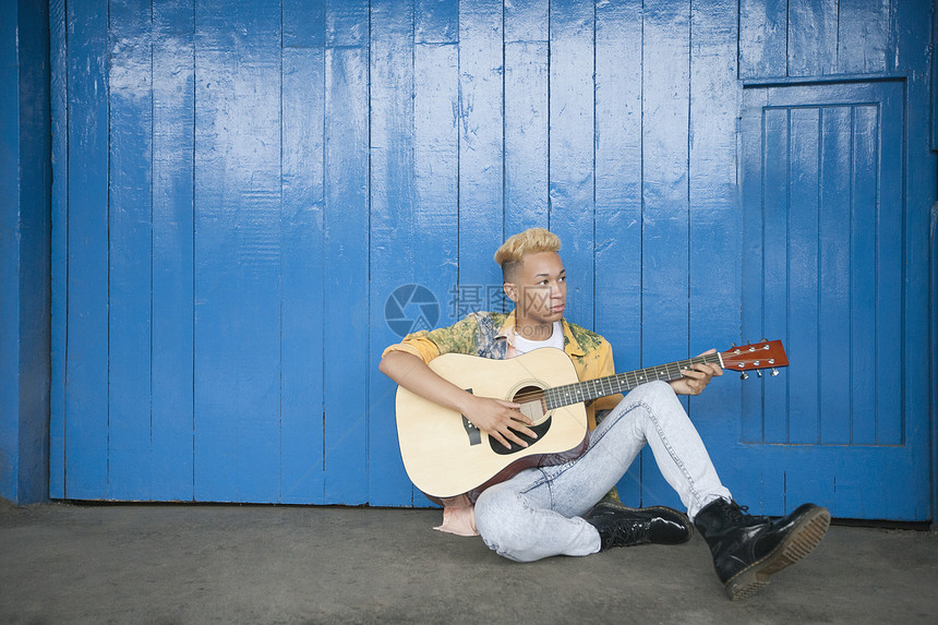 Trendy十几岁的男孩在坐在木墙壁对面时弹吉他图片