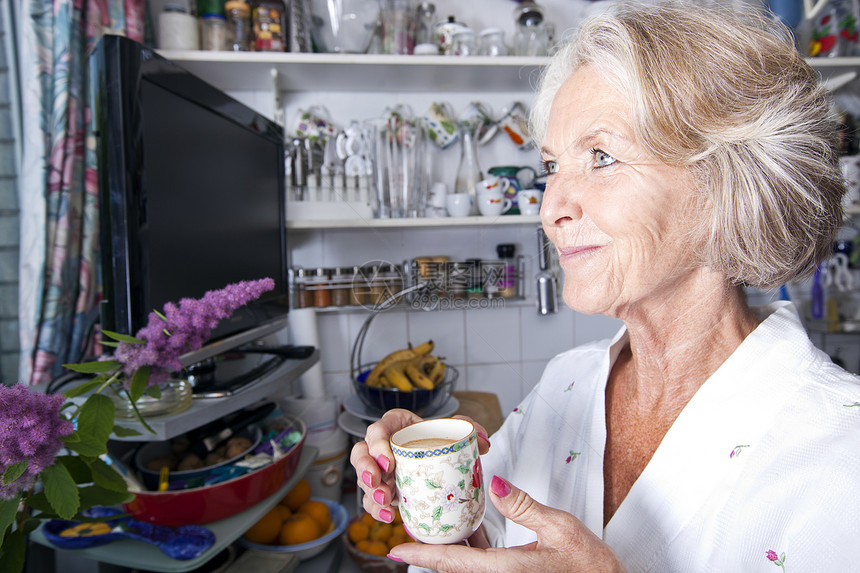 深思熟虑的老年女人在厨房里拿着咖啡杯时向外看图片