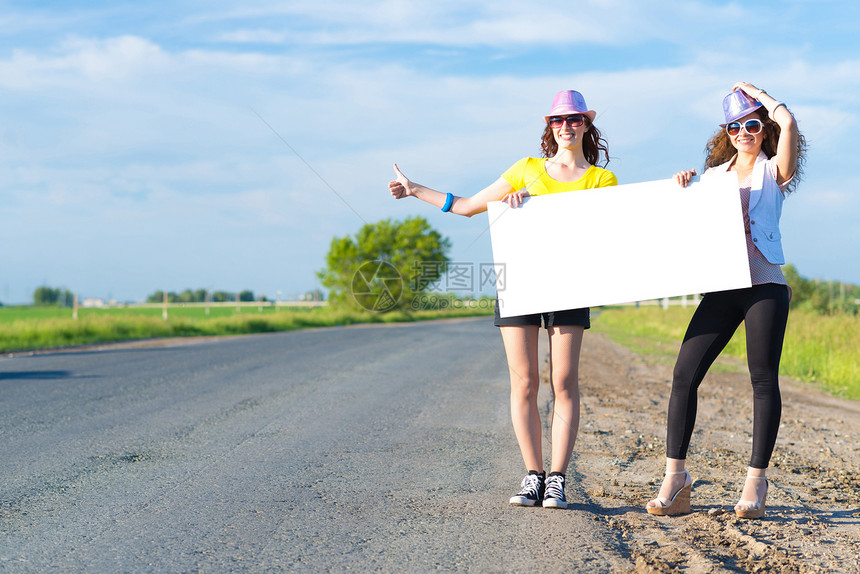 两名年轻女性站在空白的横幅上站着航程女孩冒险旅行纸板假期便车旅游女士木板图片