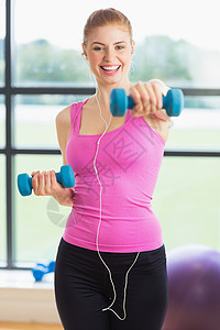 健身工作室中适合运动的女子在健身房用哑铃锻炼音乐女性力量调子训练耳机肌肉金发背心女士背景图片