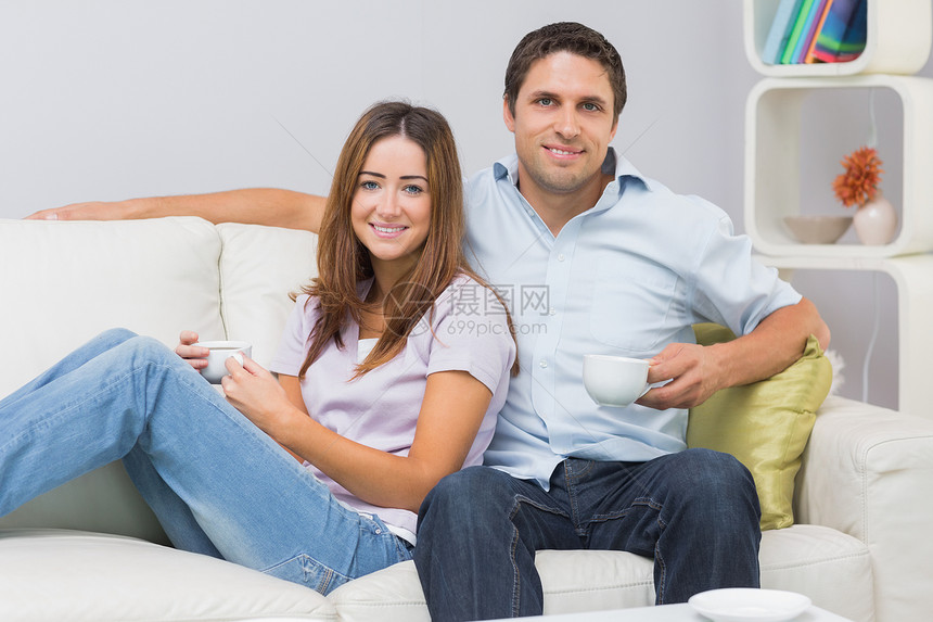 坐在沙发上和家里茶杯一起坐着的可爱情侣男性杯子客厅家庭家庭生活饮料公寓棕色长椅牛仔裤图片