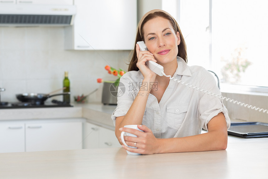 带咖啡杯的微笑女人在厨房里使用固定电话女士房子电话杯子呼唤微笑沟通女性闲暇棕色图片