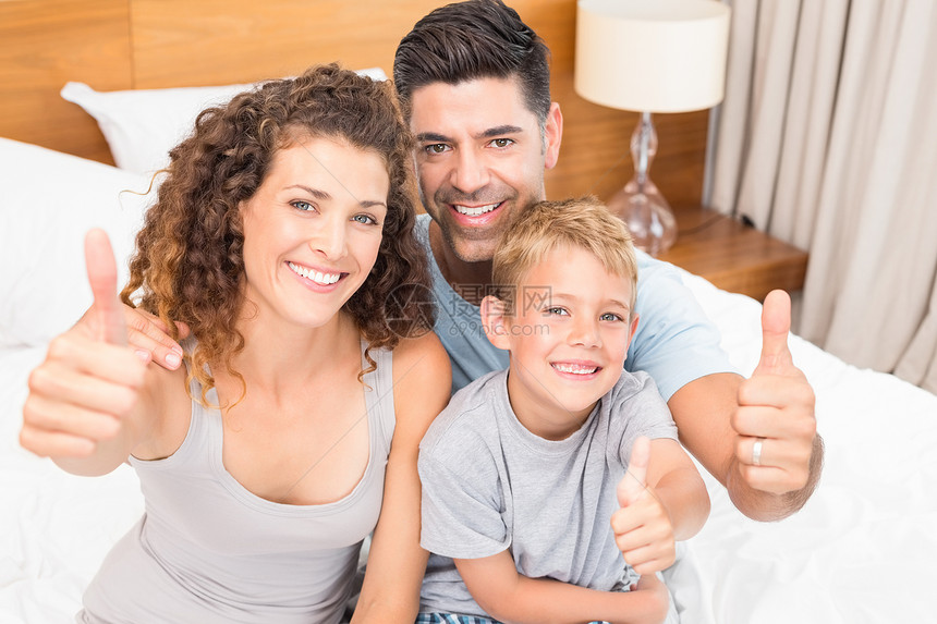 快乐的年轻家庭在床上用照相机微笑 举起拇指图片
