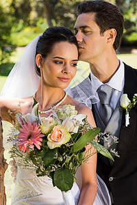 Groom在公园亲吻他的漂亮新娘女性夫妻女士妻子男人花束新郎亲热庆典团结裙子高清图片素材