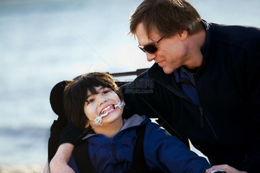 父亲与残疾儿子一起坐在湖岸边微笑轮椅照顾者多样性海洋乐趣男人家庭混血需求图片
