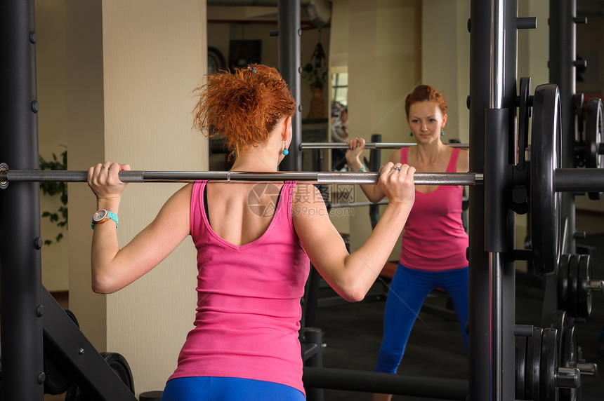 年轻女孩用巴铃做蹲地女性下蹲镜子女士健身房反射机器运动头发红色图片