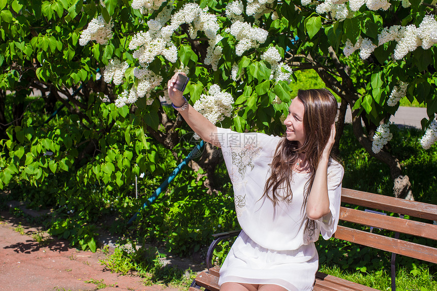 年轻女子在花开的Lilac花园用手机拍照女性公园消息享受细胞紫丁香树叶女孩花朵照片图片