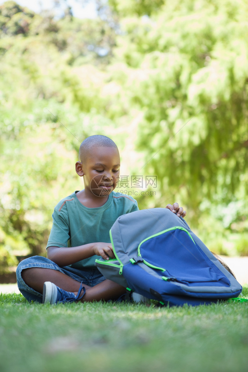 快乐的学校男孩打开他的书包坐在草地上图片