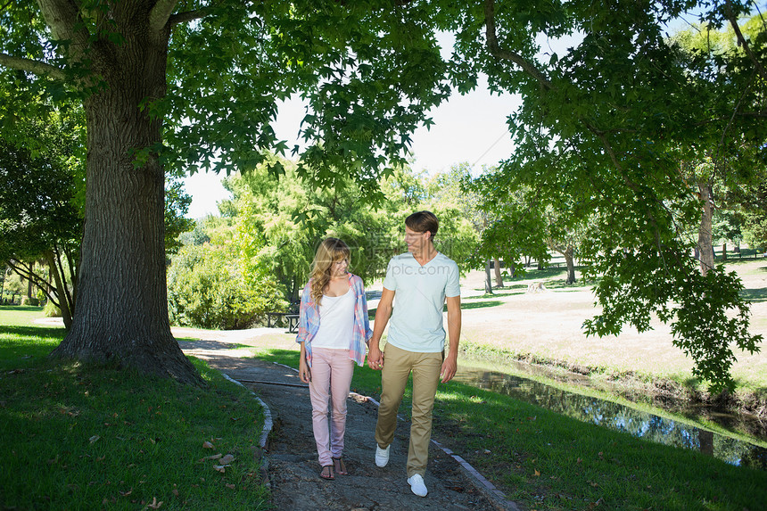 可爱的情侣在公园里手牵手行走途径衬衫女士蓝天亲密感微笑女朋友太阳绿地男朋友图片