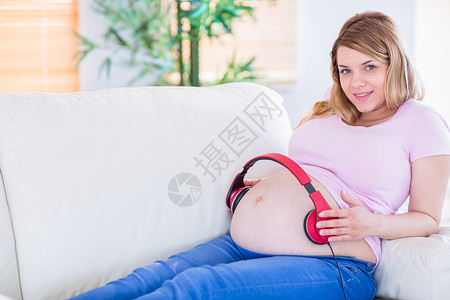微笑的怀孕妇女把耳机放在肚子上腹部育儿成人福利家庭客厅公寓女士女性长椅家庭生活高清图片素材