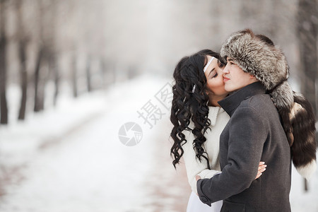 异国情调的亚洲新娘和新郎在白雪皑皑的冬季小巷中接吻 年轻人穿着外套 戴着裘皮帽 女人穿着漂亮的羊皮婚纱 寒冷季节保暖衣物 复制文背景