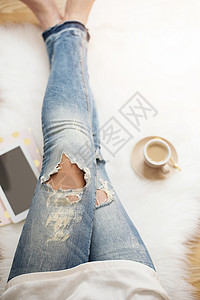 一个穿着破旧牛仔裤的年轻女子坐在家里白色毛皮地毯上的木地板上 周围一杯咖啡 平板电脑和笔记本 金色明亮的女性主题美丽的高清图片素材