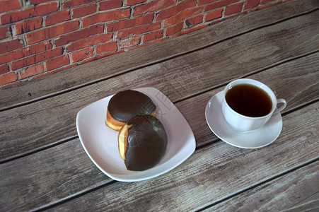 砖墙上的木桌上放着一杯放在碟子上的红茶和一个盘子 盘子里放着两个巧克力糖衣甜甜圈 特写背景墙面包杯子糕点奶油早餐飞碟油炸木头咖啡背景图片