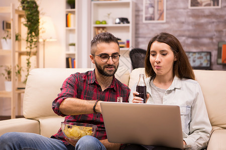 白种夫妇喝汽水和吃薯片女朋友幸福闲暇男人电脑家庭果汁沙发娱乐房子背景图片