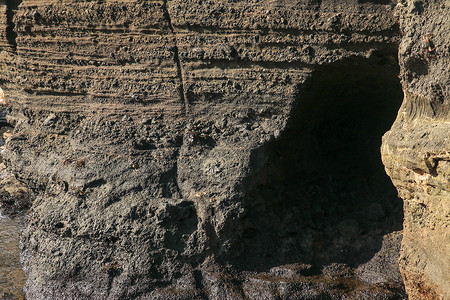 巴厘岛印度洋沿岸火山熔岩的深色岩石 一个被海浪冲进悬崖壁的小洞穴 太阳光线照亮了岩石峭壁的粗糙墙壁背景图片