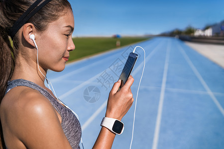 鼠年行好运跑步音乐激励女人戴着耳机听电话应用程序 赛跑者看着体育场跑道上的智能手机 耳机和手机准备好运行 健康的生活方式背景