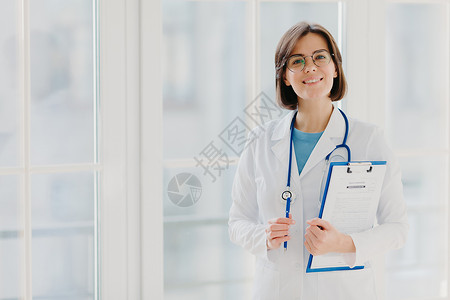 漂亮的微笑实习生拿着写有纸和笔的剪贴板 穿着白色制服 准备在自己的私人诊所看病人 站在室内白色背景下 复制空间区域背景图片