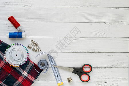 缝纫配件 白色木制背景上的格子织物 织物 缝纫线 针 别针 剪刀和缝纫厘米 顶视图 平面背景图片
