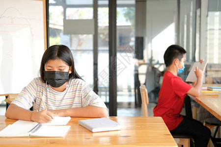 身戴面罩的女学生在做功课时写笔记学习时使用面具口罩女孩学校孩子们教育孩子家庭作业写作医疗图书馆背景图片