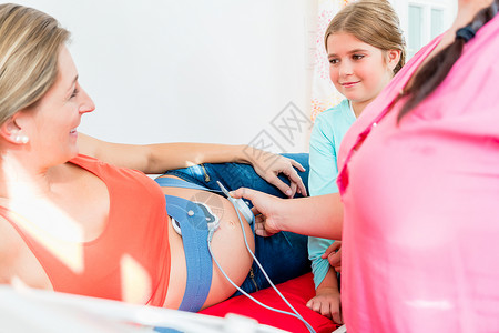 年轻女孩看助产士把CTG附在怀孕肚子上心电图产前检查心音扫描姐姐兄弟微笑后代咨询检查背景图片