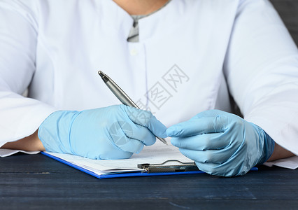 一名穿白色大衣和蓝色医疗手套的医生坐在桌子上用笔写字关心高清图片素材