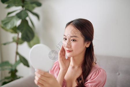年轻的亚洲女性在客厅里拿着镜子看着镜子化妆品房间女士沙发女孩光学皮肤背景图片