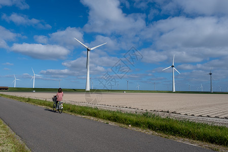弗莱风车农场旁的年轻女性电动绿色自行车 风车在美丽明亮的日子里与世隔绝 荷兰涡轮能源公园天空技术场地生态植物发电机海洋背景