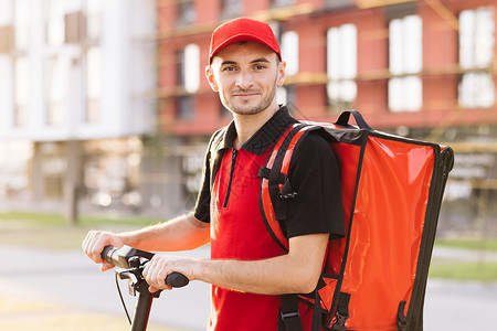 可移动板车身穿红色制服的男送货员站在街上 面对镜头的肖像照片 背着红色背包和电动滑板车的男快递员 打造绿色交通背景