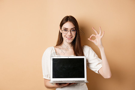 澳门回归20周年宣传展板电子商务 戴着眼镜的微笑的年轻女性显示出好的标志和笔记本电脑屏幕推荐米色背景上的互联网宣传周年工作工作室伙伴化妆品女士褐色购物女背景