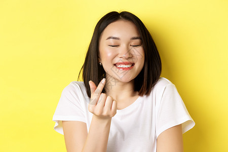 情人节和女性概念 身穿白色 T 恤的漂亮亚洲女孩的特写 微笑着展示指心 我爱你的手势 站在黄色背景上发型理发学生促销成人商业交易背景图片