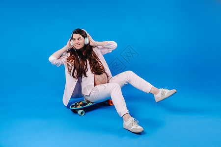 滑板爱好者穿着粉红色西装的孕妇坐在滑板上 手持耳机 在蓝色背景下听音乐活动爱好音乐享受套装木板唱歌女士工作室成人背景