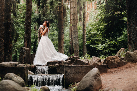 婚纱瀑布一位身着白色连衣裙 戴着手套 手捧花束的优雅新娘站在森林的溪流旁 享受着大自然 自然公园里穿着婚纱和手套的模特 白俄罗斯金发女郎背景