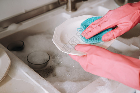 手托盘子戴粉红色手套的家庭主妇女孩用洗涤剂在水槽里用手洗碗 女孩在家里打扫房子 戴着手套洗碗盘子海绵眼镜家务台面洗涤菜肴女士公寓卫生背景