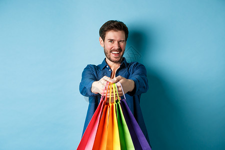 洋洋洋得意的英俊男人在闪耀和微笑 用色彩多彩的购物袋伸出双手 站在蓝背景上生活胡子促销广告购物工作室购物者情绪成功商业背景图片