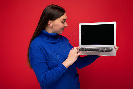照片中美丽的年轻黑头发女人拿着电脑笔记本电脑 看着蓝毛衣中的网书键盘 在红墙背景上被孤立背景图片