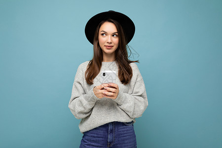 美丽的黑发年轻女性戴着黑色帽子和灰色毛衣 拿着智能手机 看着背景中孤立的侧面发短信消息冲浪电话用户互联网短信网络神器机动性背景图片