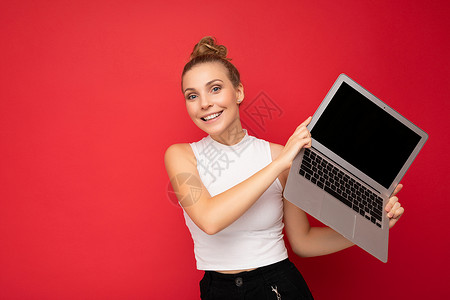 美丽微笑 迷人 快乐的金发年轻女子 头发聚集 看着相机 手持电脑笔记本电脑 身穿白色 T 恤 与红墙背景隔离互联网广告社交网页网背景图片
