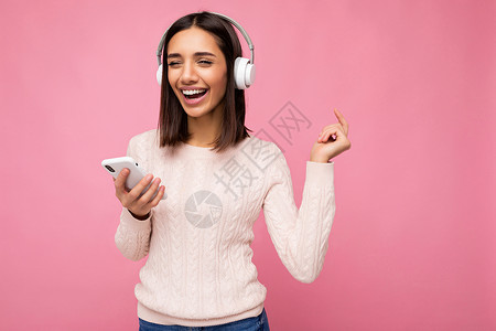 收边条照片中 美丽快乐 微笑的年轻女性穿着时尚的休闲服 与背景墙隔开 手持和使用手机 戴着白色蓝牙耳机听音乐 边看边玩歌曲互联网神器收背景