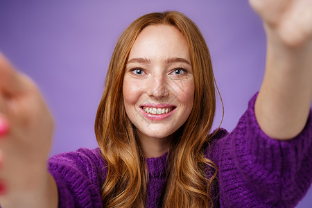 穿着紫色毛衣 真诚开朗 开朗的姜女 手伸着相机 用友好乐观的目光微笑着 自拍或想在紫色墙上拿东西背景图片