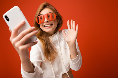 迷人迷人的年轻微笑快乐的女人手持和使用手机自拍 穿着时髦的衣服 与墙壁背景隔离眼镜社交互联网神器博主电话金发女郎白色摄影帽衫背景图片