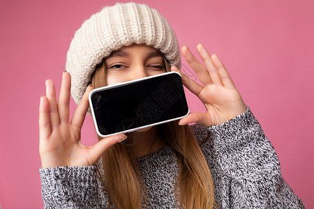 眨眼的特写照片美丽的年轻金发女子穿着休闲灰色毛衣和米色帽子 在粉红色背景中被隔离 手里拿着 并展示手机与空显示屏 用于模拟看相机背景图片