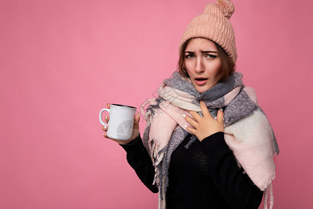 穿着黑色毛衣帽和温暖围巾的迷人而生病的年轻黑头发女性男子 在粉红背景上被孤立 手持纸杯 用于喝热茶 并观看有冷 感冒的照相机热可可高清图片素材