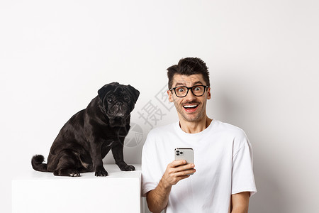 带着可爱的黑小狗坐着 使用手机 站在白色背景上 Y I L K 我宠物广告男性微笑博主犬类行动横幅电话胡须背景图片