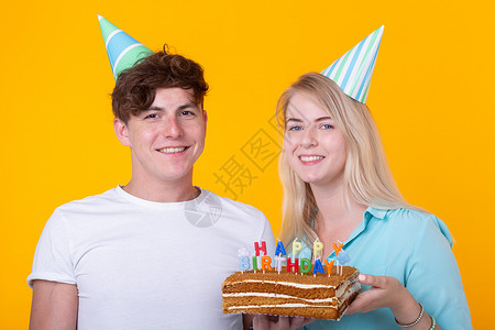 有趣的年轻夫妇戴着纸帽 拿着蛋糕 站在黄色背景下 做着傻脸 祝生日快乐 祝贺和鬼混的概念礼物派对创造力蓝色节日惊喜装饰风格情绪周背景图片