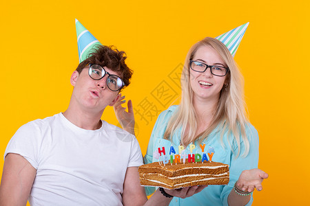 有趣的年轻夫妇戴着纸帽 拿着蛋糕 站在黄色背景下 做着傻脸 祝生日快乐 祝贺和鬼混的概念乐趣派对甜点庆典工作室喜悦海报娱乐潮人节背景图片