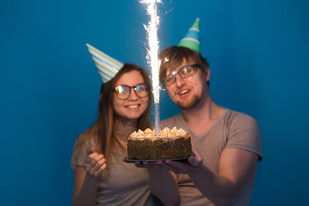 有趣的年轻夫妇戴着纸帽 拿着蛋糕 站在蓝色背景下 做出一张傻脸 祝生日快乐 祝贺和鬼混的概念艺术情绪娱乐风格惊喜海报周年派对创造背景图片