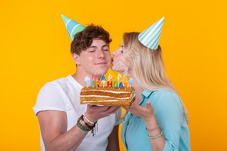 有趣的年轻夫妇戴着纸帽 拿着蛋糕 站在黄色背景下 做着傻脸 祝生日快乐 祝贺和鬼混的概念娱乐庆典潮人礼物喜悦蓝色海报乐趣纪念日惊背景图片