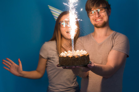 有趣的年轻夫妇戴着纸帽 拿着蛋糕 站在蓝色背景下 做出一张傻脸 祝生日快乐 祝贺和鬼混的概念纪念日娱乐节日生日女孩甜点艺术礼物创背景图片